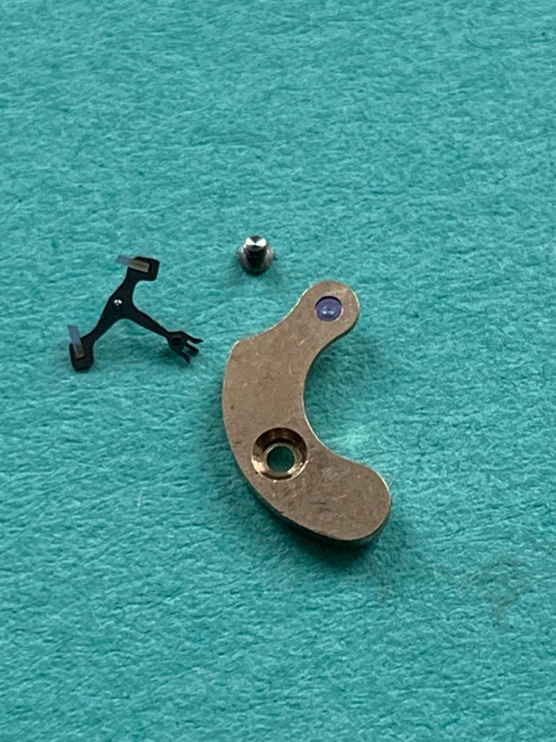 May include: A collection of watch parts, including a small screw, a curved brass-colored lever with a hole, and a black metal component with multiple prongs. The parts are arranged on a teal-colored surface.