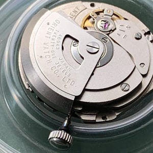 May include: A detailed view of a watch movement, showcasing intricate gears and parts. The silver mechanism displays the text "ORIENT WATCH" and "JAPAN JEWELS CO. LTD." The watch crown is visible. The movement is housed within a clear container.