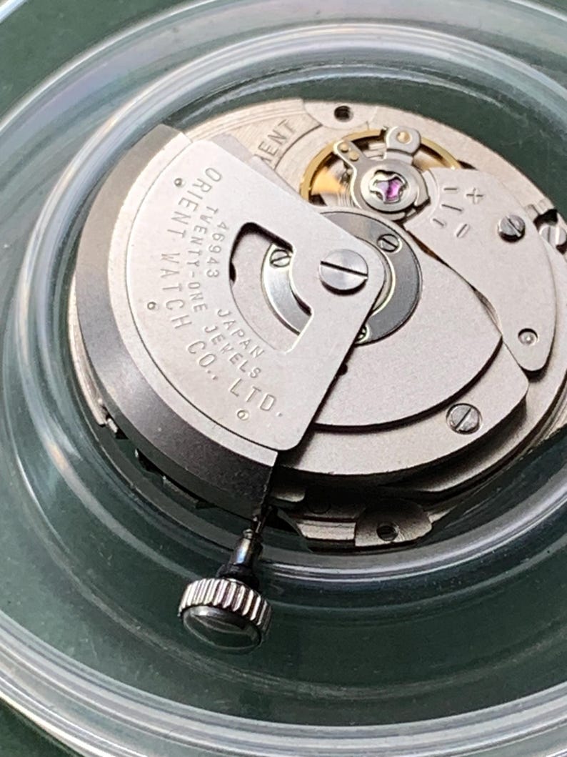 May include: A detailed view of a watch movement, showcasing intricate gears and parts. The silver mechanism displays the text "ORIENT WATCH" and "JAPAN JEWELS CO. LTD." The watch crown is visible. The movement is housed within a clear container.