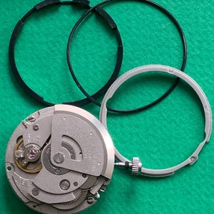 May include: A disassembled watch movement with a silver-colored mechanism, a gray bezel, and two black rubber rings. The watch components are arranged on a green surface, showcasing the intricate internal workings of the timepiece.