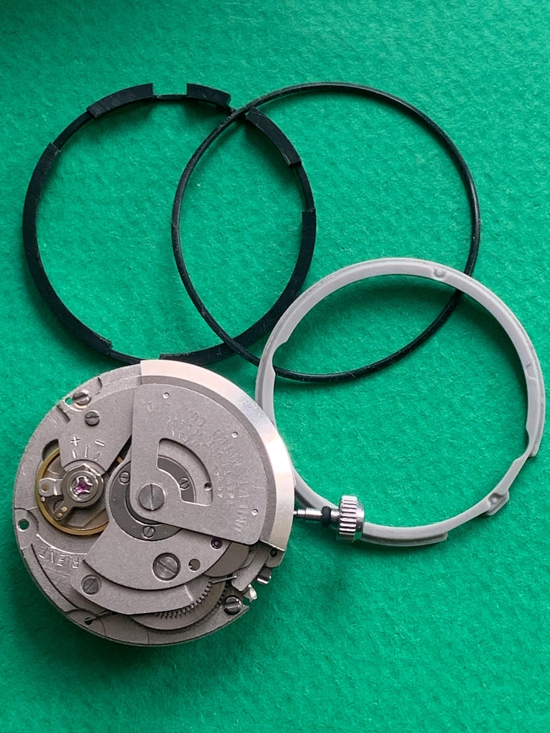 May include: A disassembled watch movement with a silver-colored mechanism, a gray bezel, and two black rubber rings. The watch components are arranged on a green surface, showcasing the intricate internal workings of the timepiece.