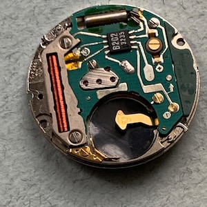 May include: Close-up of a watch movement with a green circuit board, metallic components, and a black battery. The board has the text "B202 3235". The movement is circular and has various electronic components.