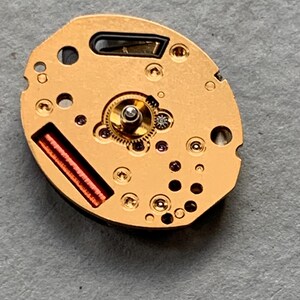 May include: A gold-coloured watch movement, featuring detailed gears, tiny jewels, and a red coil. The oval mechanism has several holes and is set against a grey background, highlighting its intricate design and craftsmanship.