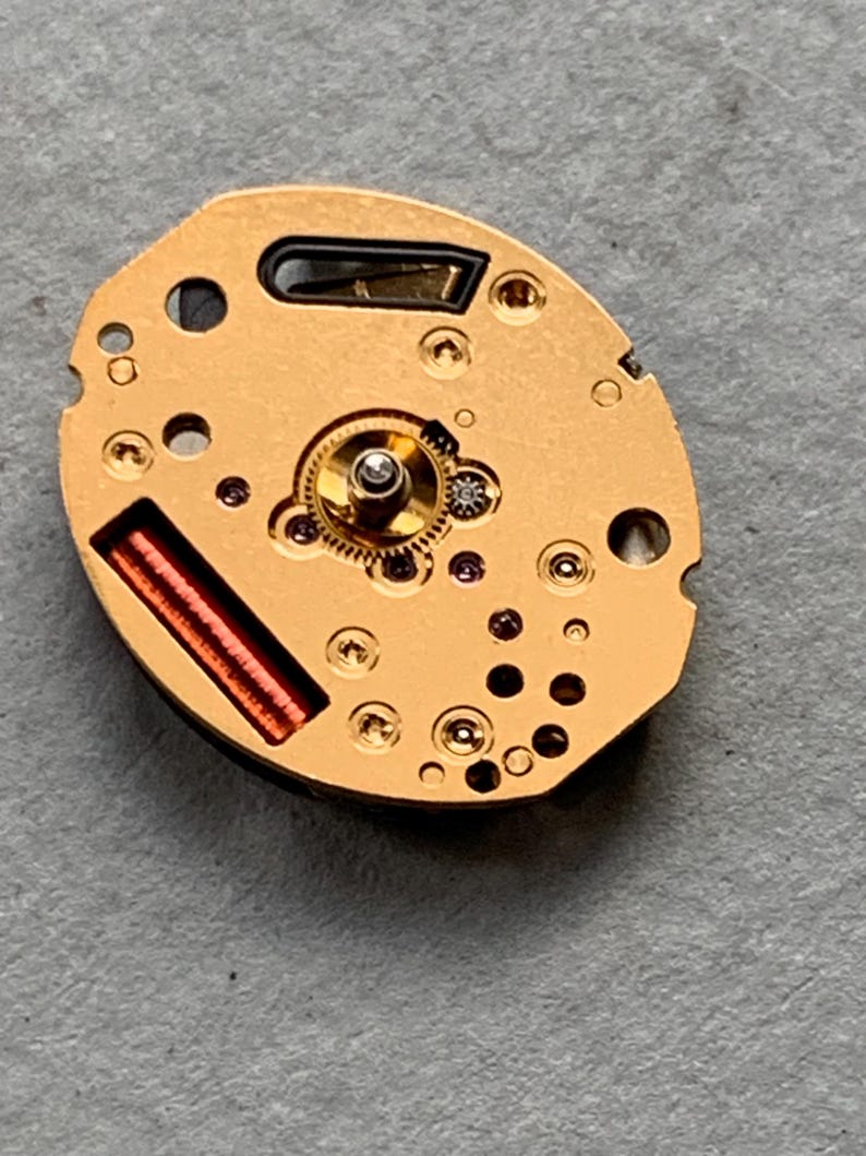May include: A gold-coloured watch movement, featuring detailed gears, tiny jewels, and a red coil. The oval mechanism has several holes and is set against a grey background, highlighting its intricate design and craftsmanship.