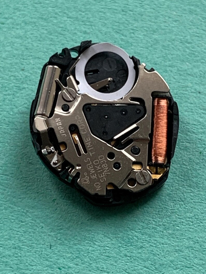 May include: Close-up of a watch movement with intricate mechanical components. The movement features silver, copper, and black elements, including a coil, gears, and the text "NO JEWELS SEIKO TIME CORP 7N83D JAPAN".