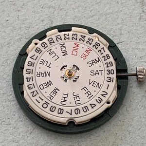 May include: Close-up of a watch movement with a white dial and a dark green frame. The dial displays the days of the week in French and numbers 1-31. A small metal crown is visible on the right side.