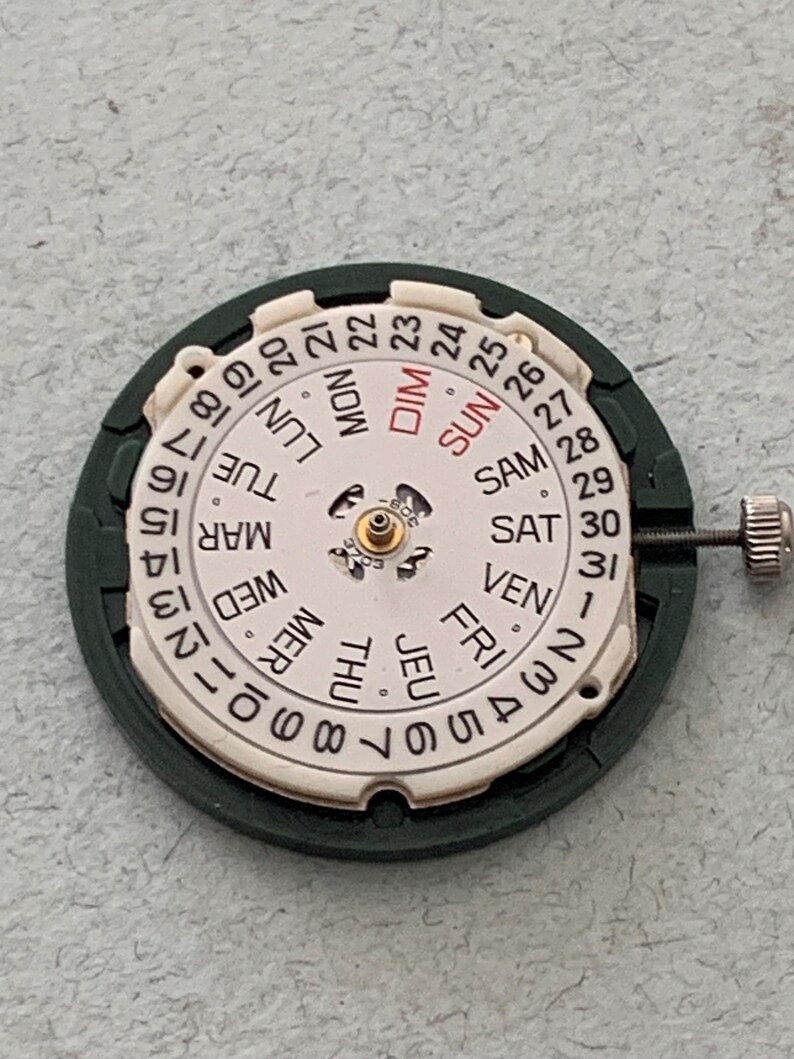 May include: Close-up of a watch movement with a white dial and a dark green frame. The dial displays the days of the week in French and numbers 1-31. A small metal crown is visible on the right side.