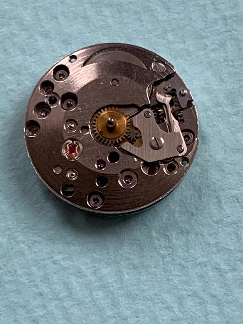May include: A close-up of a silver watch movement with a gold gear and several small screws.