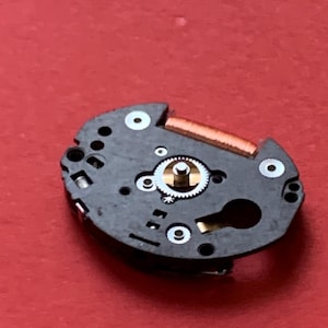 May include: A close-up of a watch movement on a red surface. The movement is black with gold and silver components, including gears, a balance wheel, and a copper-colored coil. Several white circles are visible.