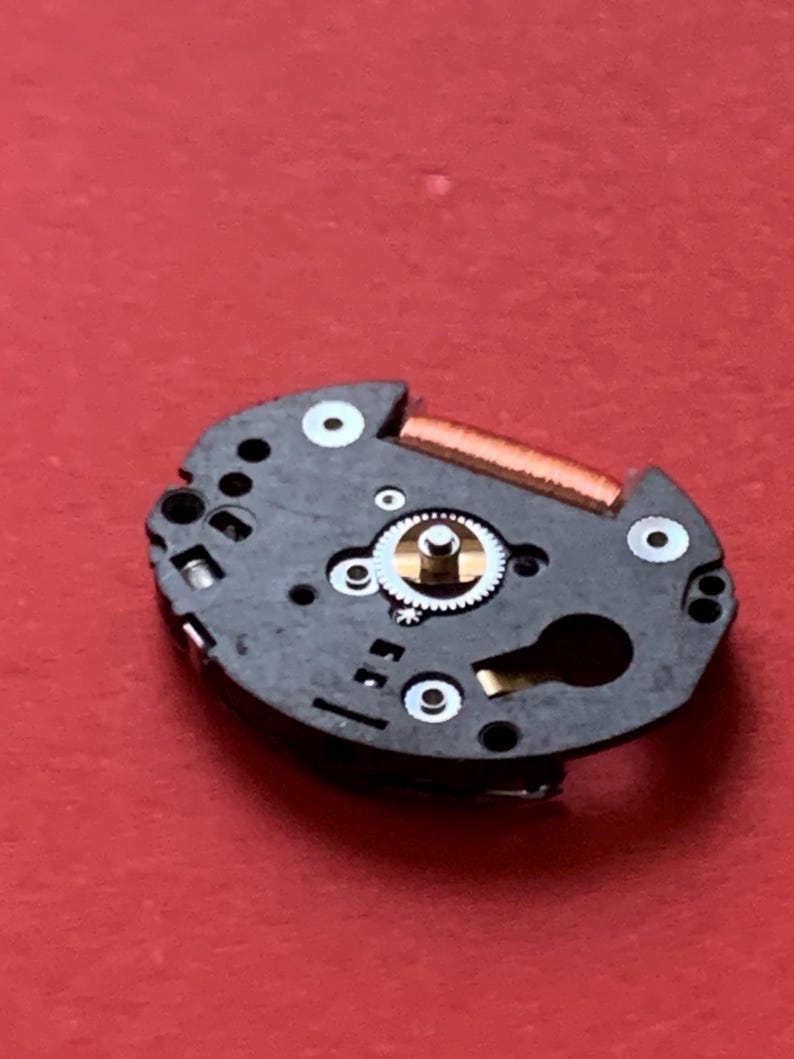 May include: A close-up of a watch movement on a red surface. The movement is black with gold and silver components, including gears, a balance wheel, and a copper-colored coil. Several white circles are visible.