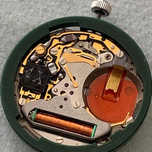 May include: Close-up of a Citizen watch movement, model 7302A, with a green outer casing. The internal components include gold-colored gears, a red battery, and a green and copper coil. The watch mechanism is designed for precision and durability.