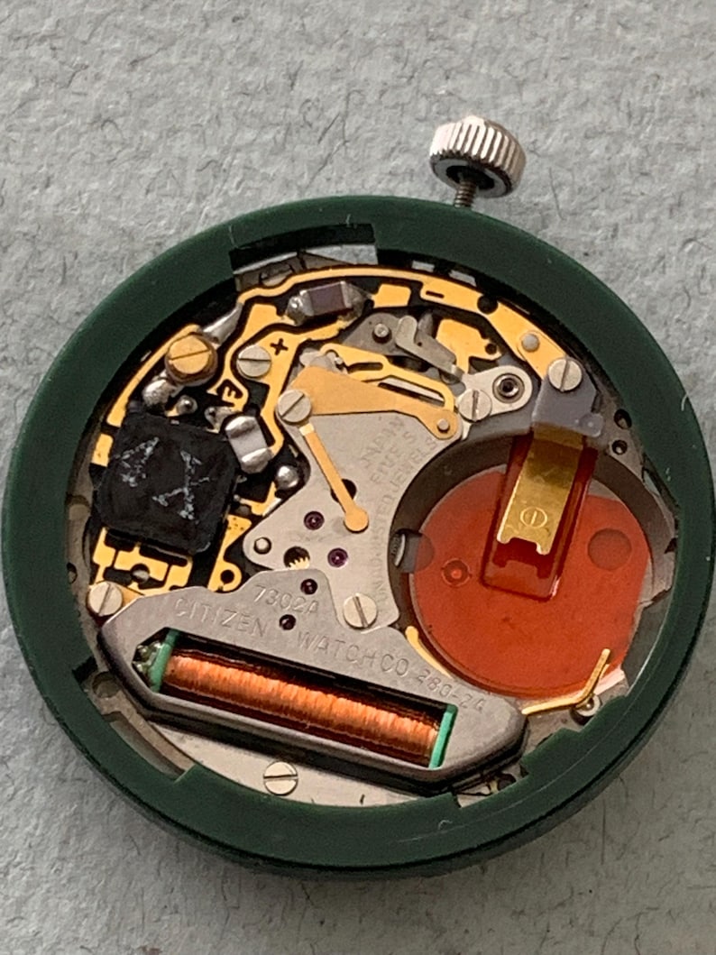 May include: Close-up of a Citizen watch movement, model 7302A, with a green outer casing. The internal components include gold-colored gears, a red battery, and a green and copper coil. The watch mechanism is designed for precision and durability.