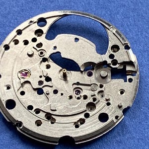 Puede incluir: Primer plano de un movimiento de reloj plateado con intrincados engranajes, agujeros y peque&ntilde;as joyas moradas. El mecanismo circular se encuentra sobre un fondo azul, mostrando la detallada artesan&iacute;a de los componentes internos del reloj.