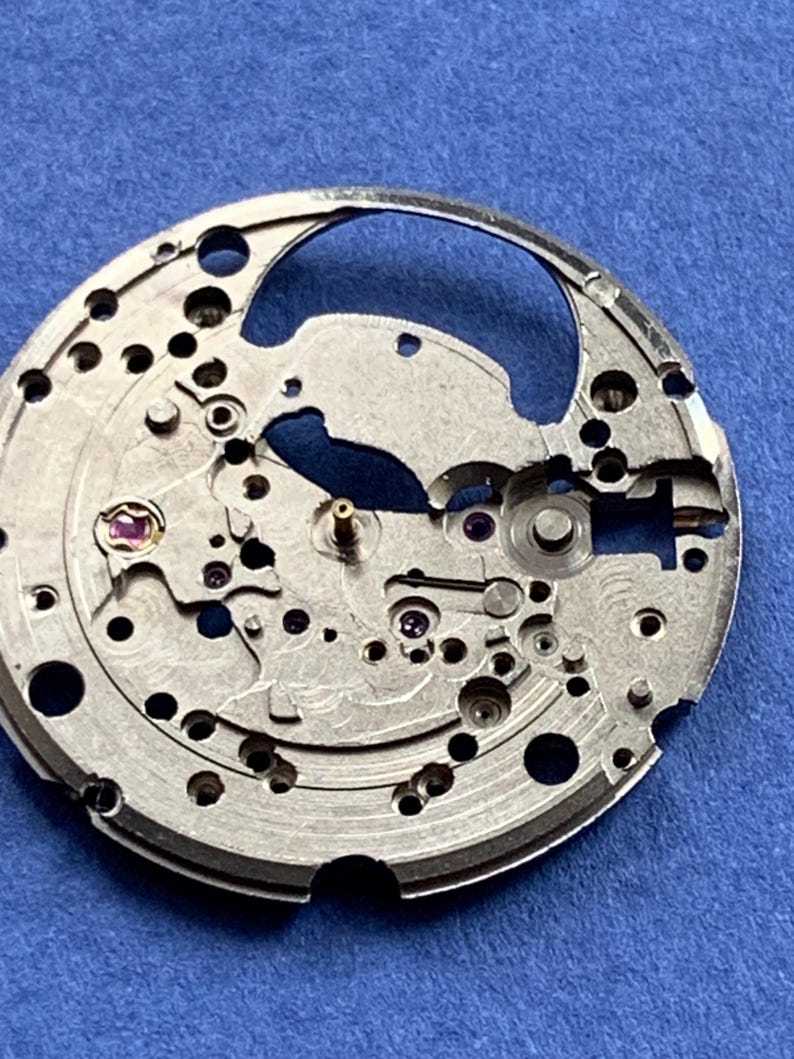 Puede incluir: Primer plano de un movimiento de reloj plateado con intrincados engranajes, agujeros y peque&ntilde;as joyas moradas. El mecanismo circular se encuentra sobre un fondo azul, mostrando la detallada artesan&iacute;a de los componentes internos del reloj.