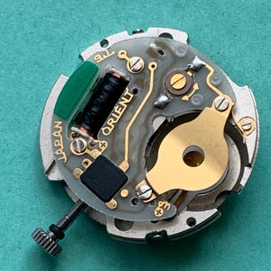 May include: Close-up of a watch movement with the brand name "ORIENT" visible. The movement features gold-colored components, a black coil, and a green rectangular piece with numbers. The words "JAPAN" and "52350" are also visible.