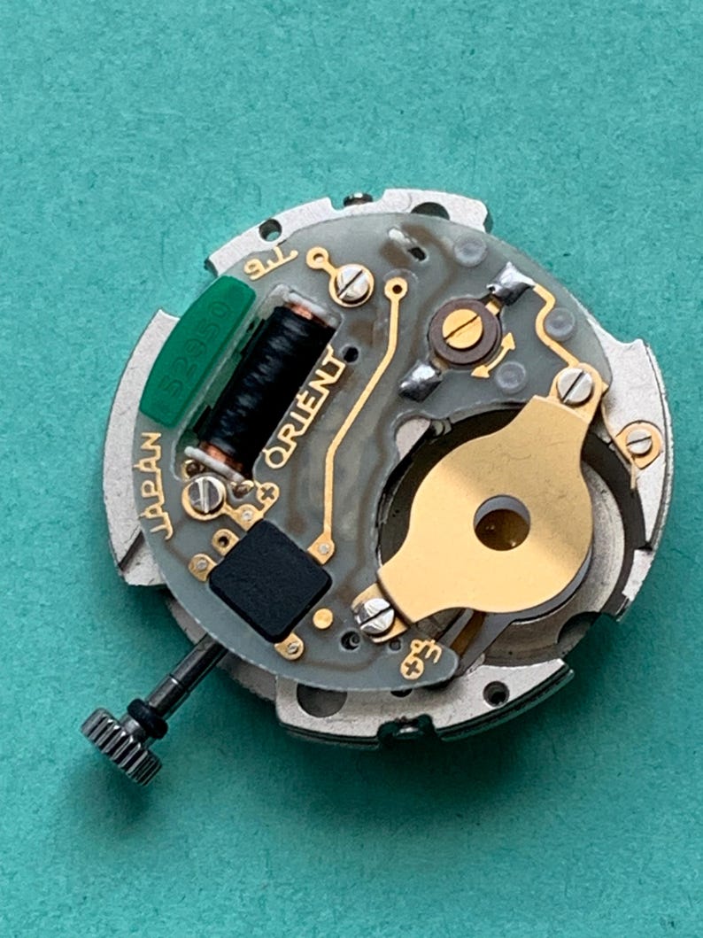 May include: Close-up of a watch movement with the brand name "ORIENT" visible. The movement features gold-colored components, a black coil, and a green rectangular piece with numbers. The words "JAPAN" and "52350" are also visible.