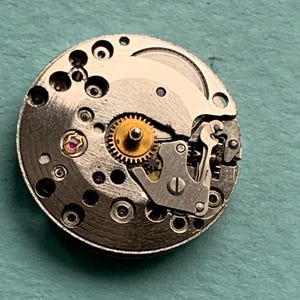 May include: A close-up of a silver watch movement with visible gears and a small pink jewel.