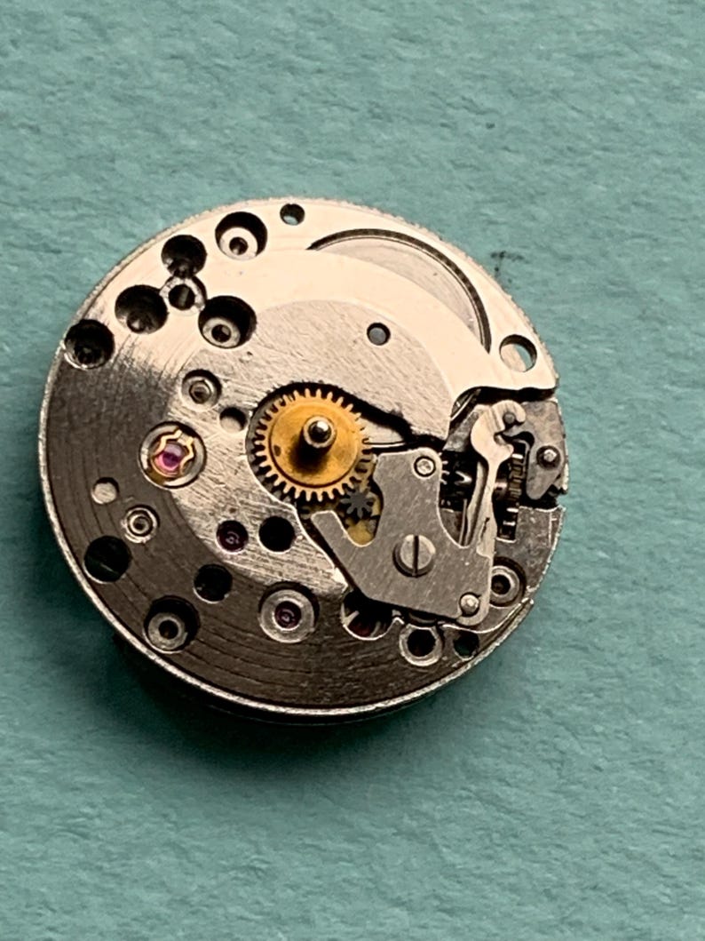 May include: A close-up of a silver watch movement with visible gears and a small pink jewel.