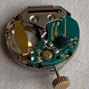 Puede incluir: Primer plano de un movimiento de reloj con una placa de circuito verde y componentes dorados. Se puede ver el texto "UNADJUSTED" y "SEVEN 7 JEWELS SWISS". Tambi&eacute;n hay una corona dorada.