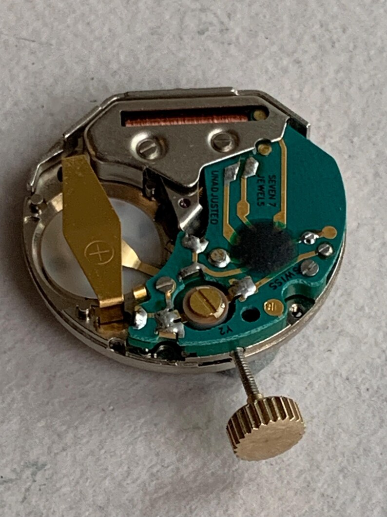 Puede incluir: Primer plano de un movimiento de reloj con una placa de circuito verde y componentes dorados. Se puede ver el texto "UNADJUSTED" y "SEVEN 7 JEWELS SWISS". Tambi&eacute;n hay una corona dorada.