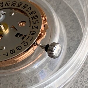 Puede incluir: Primer plano de un movimiento de reloj con una corona plateada y un dial de fecha. El dial presenta n&uacute;meros negros sobre un fondo plateado, con un anillo dorado. La corona es estriada y tiene un sello de goma negro. Las piezas del reloj est&aacute;n dentro de un recipiente de pl&aacute;stico transparente.