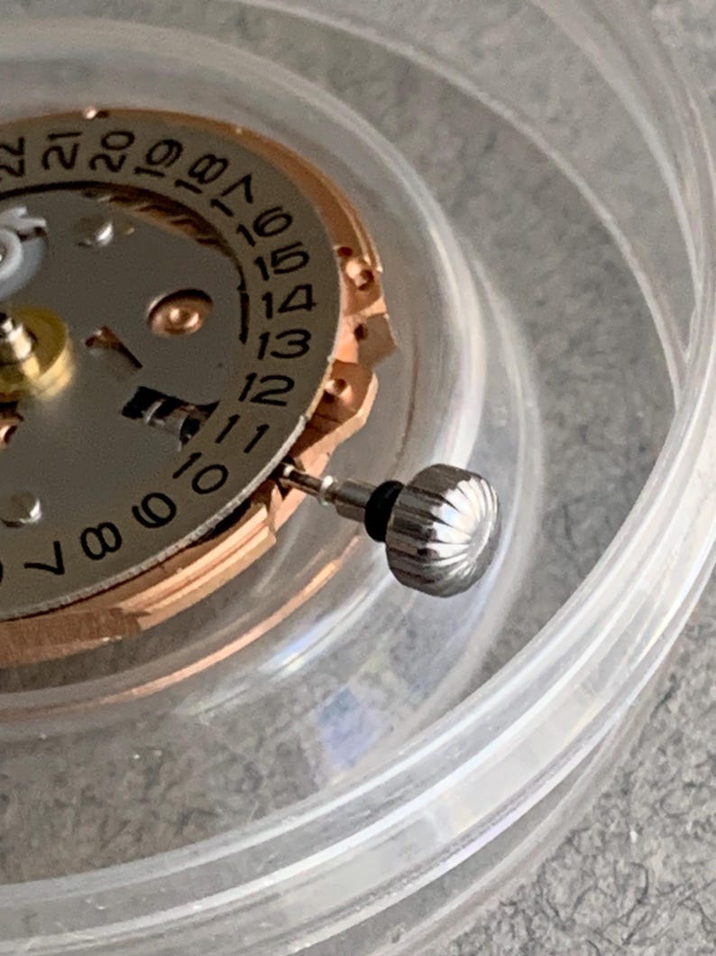 Puede incluir: Primer plano de un movimiento de reloj con una corona plateada y un dial de fecha. El dial presenta n&uacute;meros negros sobre un fondo plateado, con un anillo dorado. La corona es estriada y tiene un sello de goma negro. Las piezas del reloj est&aacute;n dentro de un recipiente de pl&aacute;stico transparente.