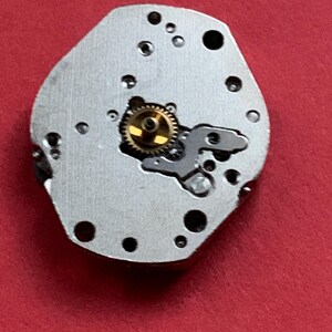 May include: A close-up of a watch movement component. The silver-colored piece has several small holes and a gold-colored gear in the center. The background is a solid red.