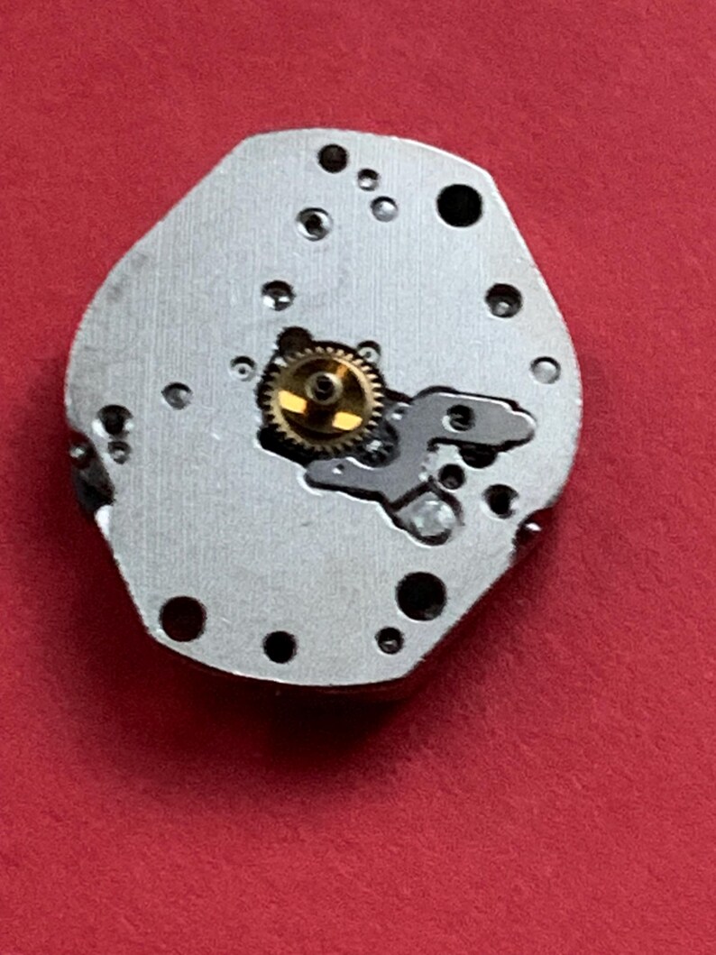 May include: A close-up of a watch movement component. The silver-colored piece has several small holes and a gold-colored gear in the center. The background is a solid red.