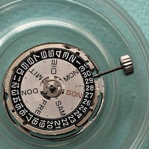 May include: A watch movement with a silver and black dial, displaying the days of the week in English and numbers for the date. The dial is surrounded by a silver metal ring and a clear plastic container.