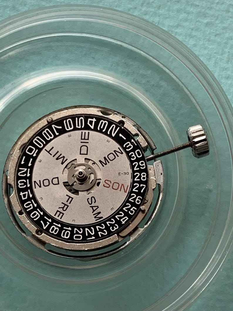 May include: A watch movement with a silver and black dial, displaying the days of the week in English and numbers for the date. The dial is surrounded by a silver metal ring and a clear plastic container.