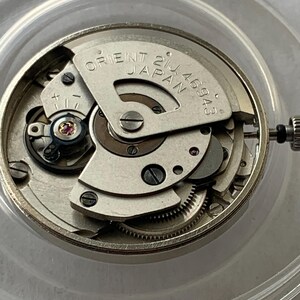 May include: Close-up of a silver Orient watch movement. The watch mechanism is visible, with intricate gears, screws, and a balance wheel. The text "ORIENT JAPAN 21J 46943" is printed on a metal plate.