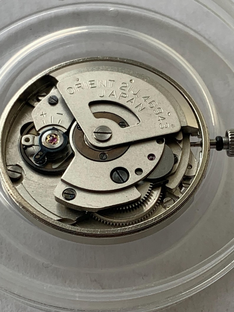 May include: Close-up of a silver Orient watch movement. The watch mechanism is visible, with intricate gears, screws, and a balance wheel. The text "ORIENT JAPAN 21J 46943" is printed on a metal plate.