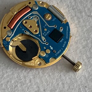 May include: Close-up of a watch movement with a gold-colored frame and a blue circuit board. The board has gold-colored components, a black square, and the text "SWISS V8 M". A screw is attached to the side. The text "UNADJUSTED SEVEN 7 JEWELS" is visible.