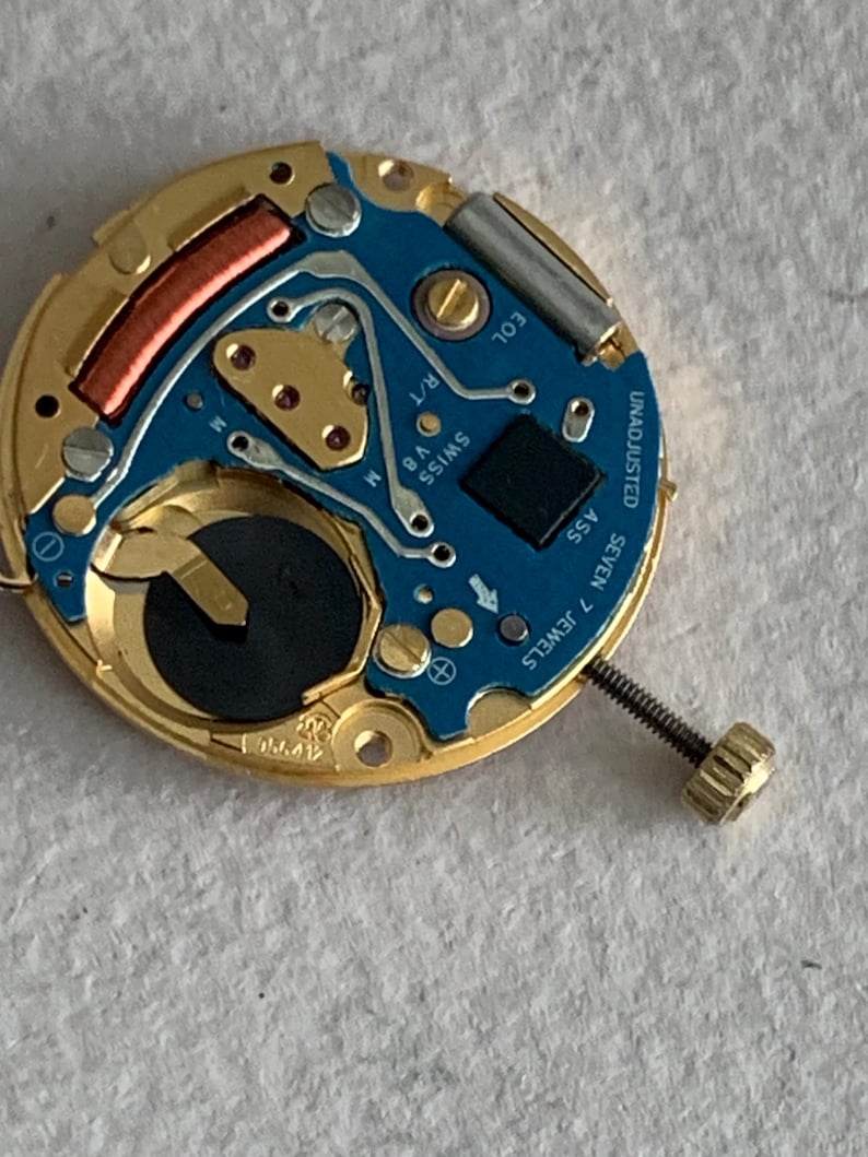 May include: Close-up of a watch movement with a gold-colored frame and a blue circuit board. The board has gold-colored components, a black square, and the text "SWISS V8 M". A screw is attached to the side. The text "UNADJUSTED SEVEN 7 JEWELS" is visible.