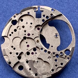 Puede incluir: Un movimiento de reloj plateado con intrincados engranajes, agujeros y un dise&ntilde;o circular. El movimiento tiene peque&ntilde;os acentos en forma de joya y est&aacute; marcado con las letras "AS" y el n&uacute;mero "1955". El fondo es azul liso.