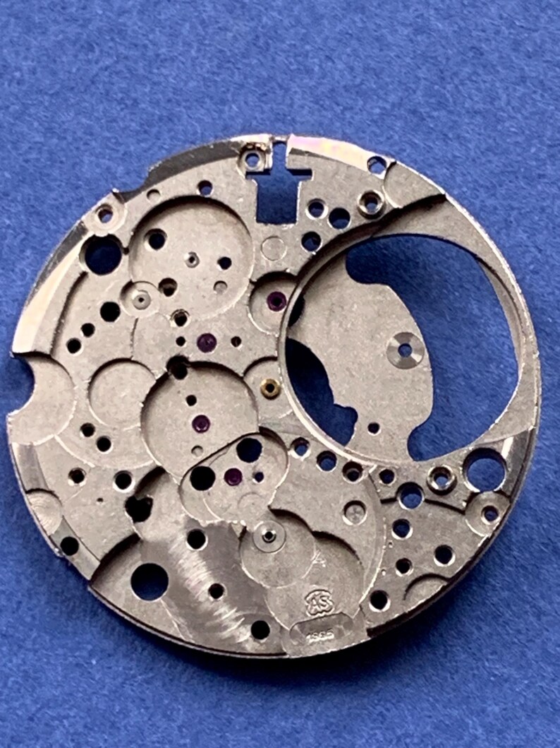 Puede incluir: Un movimiento de reloj plateado con intrincados engranajes, agujeros y un dise&ntilde;o circular. El movimiento tiene peque&ntilde;os acentos en forma de joya y est&aacute; marcado con las letras "AS" y el n&uacute;mero "1955". El fondo es azul liso.