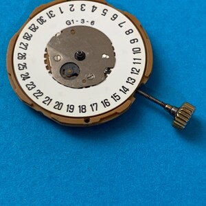 May include: A watch movement with a white dial featuring black numerals for the days of the month, from 1 to 31. The movement has a gold-colored metal casing and a small crown. The text "G1-3-6" is visible on the dial. The background is blue.