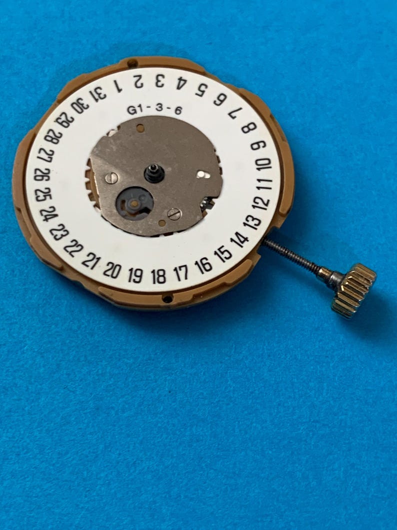 May include: A watch movement with a white dial featuring black numerals for the days of the month, from 1 to 31. The movement has a gold-colored metal casing and a small crown. The text "G1-3-6" is visible on the dial. The background is blue.