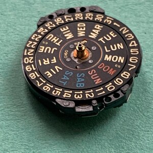 May include: A black watch calendar wheel with gold-colored text and numbers. The days of the week are labeled in multiple languages. The numbers 1-31 are displayed around the outer edge. The wheel has a central brass-colored post.