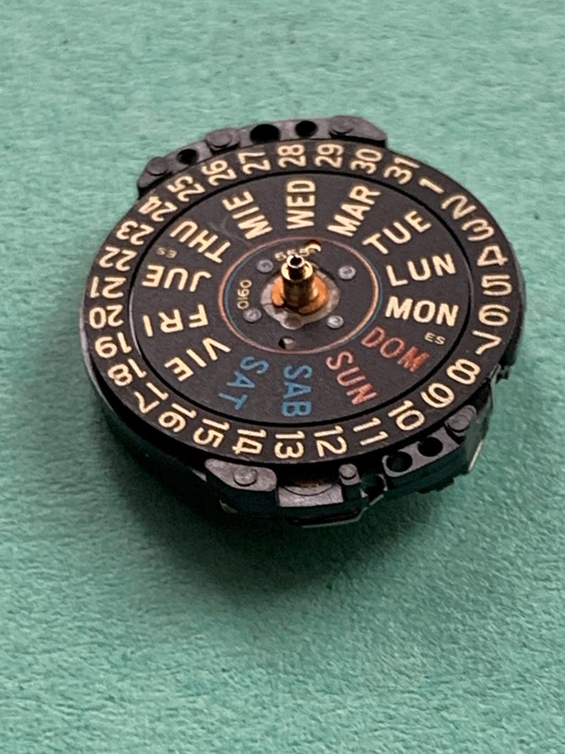 May include: A black watch calendar wheel with gold-colored text and numbers. The days of the week are labeled in multiple languages. The numbers 1-31 are displayed around the outer edge. The wheel has a central brass-colored post.