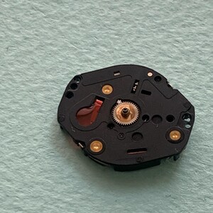 May include: Close-up of a black watch movement with gold-coloured accents. The watch mechanism features a small gear, a red component, and several gold-coloured screws. The background is a light green surface.