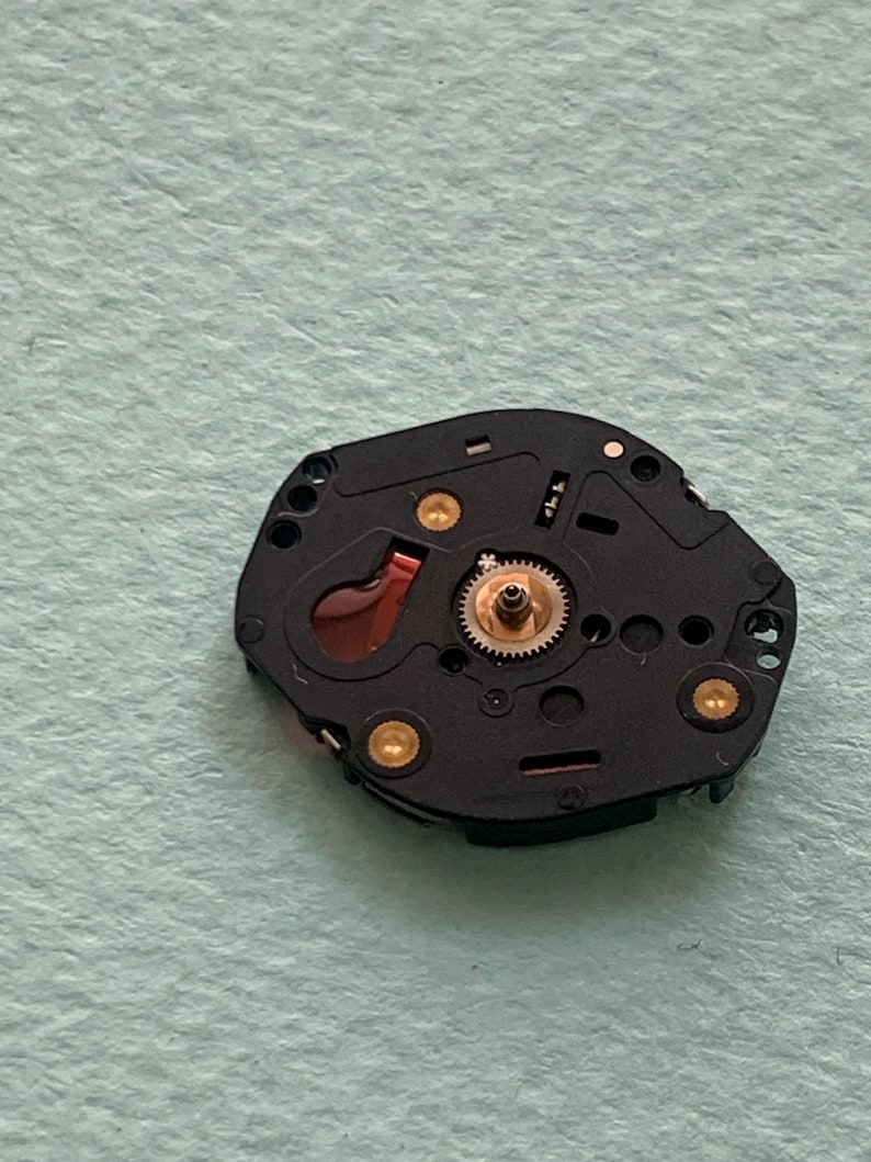 May include: Close-up of a black watch movement with gold-coloured accents. The watch mechanism features a small gear, a red component, and several gold-coloured screws. The background is a light green surface.