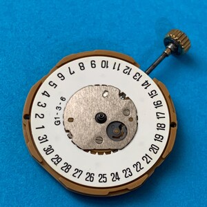 May include: A watch movement with a white dial featuring black numbers from 1 to 31, and a gold-colored outer ring. A small metal mechanism is visible in the center, with a gold-colored crown and stem extending from the side. The background is a solid blue.