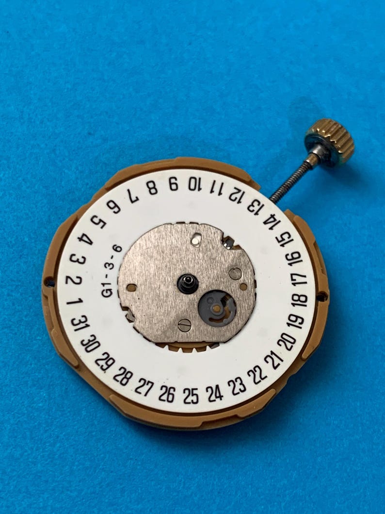 May include: A watch movement with a white dial featuring black numbers from 1 to 31, and a gold-colored outer ring. A small metal mechanism is visible in the center, with a gold-colored crown and stem extending from the side. The background is a solid blue.
