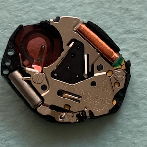 May include: A detailed view of a watch movement, showcasing intricate parts. It includes a copper coil, a red battery, and the text "SEIKO TIME CORP." The movement is set against a light blue backdrop.