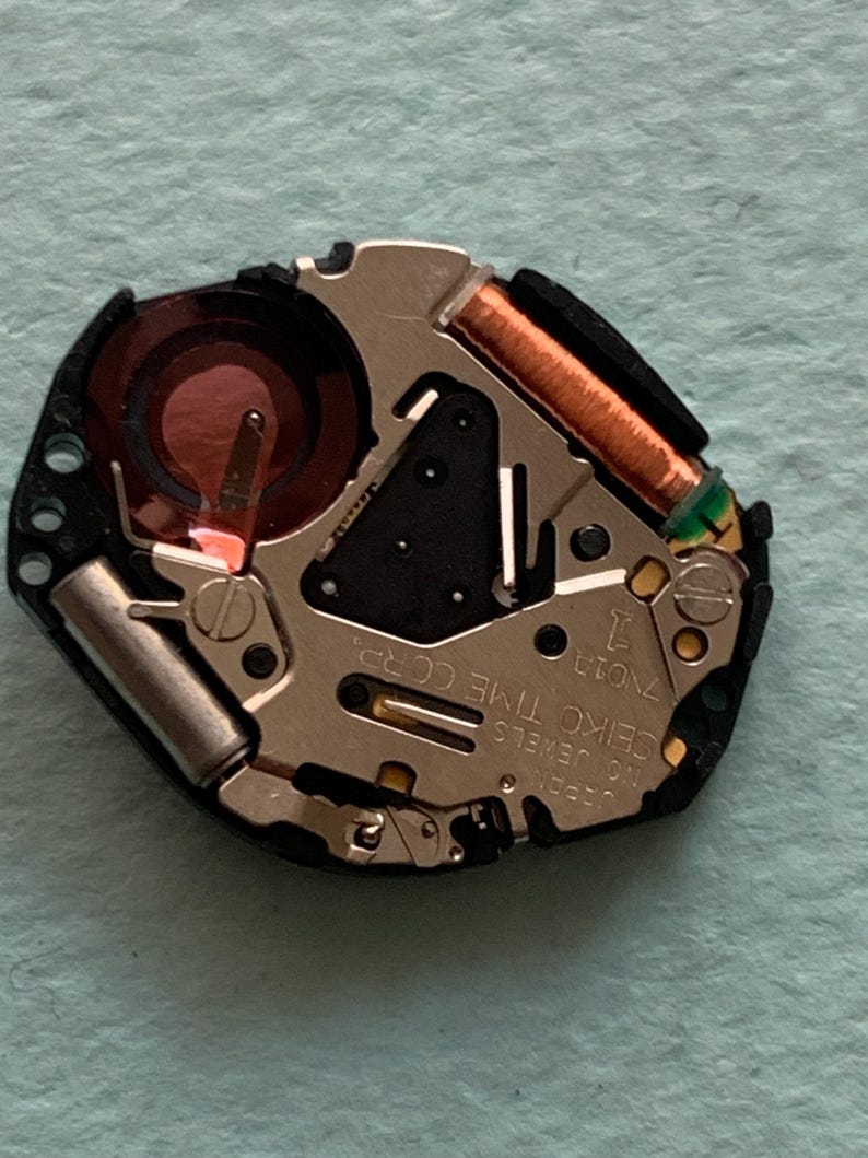 May include: A detailed view of a watch movement, showcasing intricate parts. It includes a copper coil, a red battery, and the text "SEIKO TIME CORP." The movement is set against a light blue backdrop.
