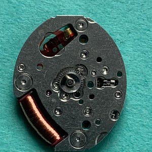May include: Close-up of a watch movement with a silver-colored base. The movement features a copper-colored coil, small gears, and various components. The background is a teal-colored surface.