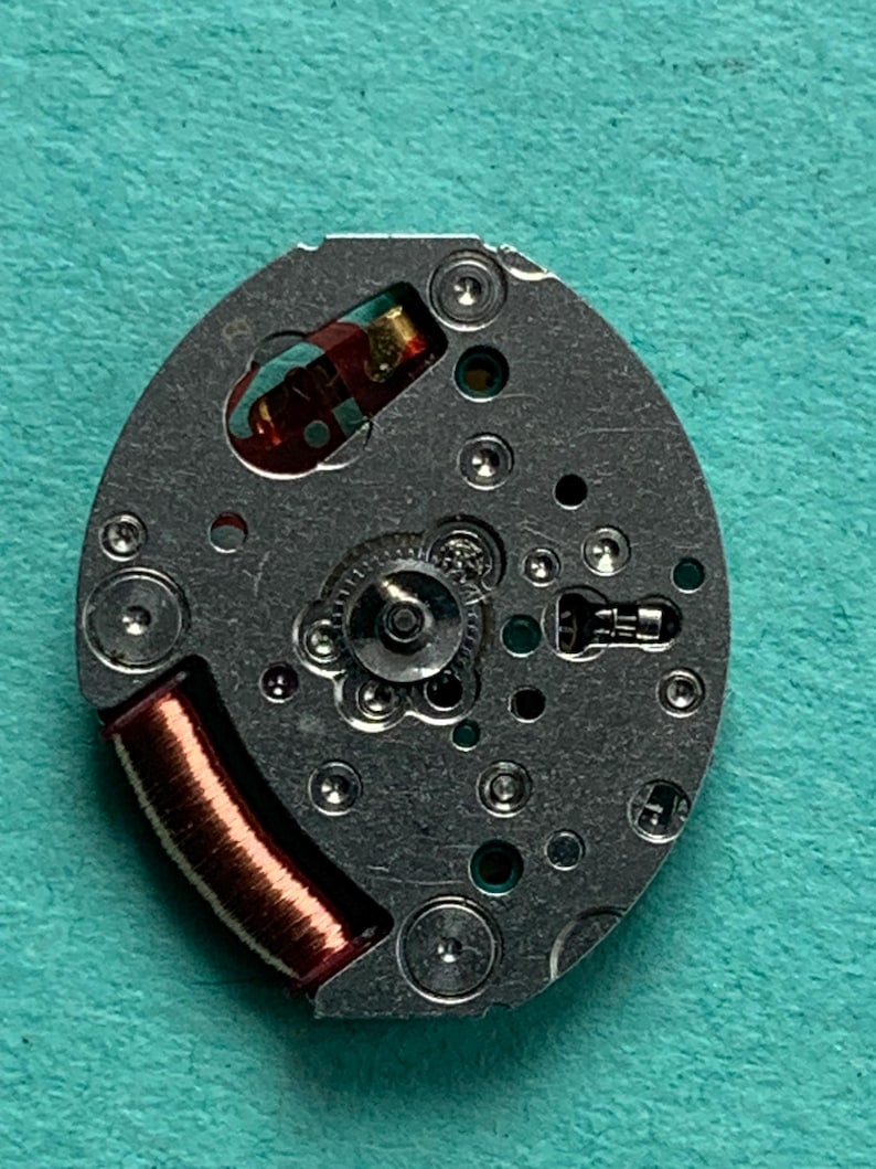 May include: Close-up of a watch movement with a silver-colored base. The movement features a copper-colored coil, small gears, and various components. The background is a teal-colored surface.