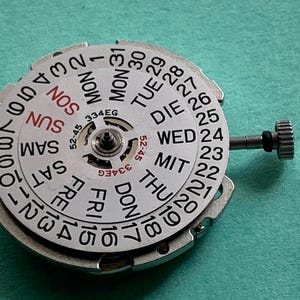 May include: A close-up of a watch movement with a white dial featuring the days of the week and numbered dates. The dial has a central mechanism and a small crown. The text "334EG" is visible.