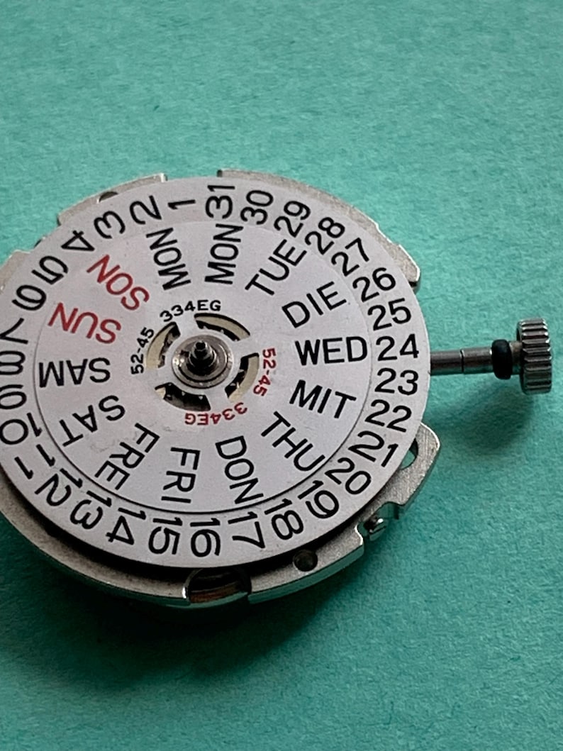 May include: A close-up of a watch movement with a white dial featuring the days of the week and numbered dates. The dial has a central mechanism and a small crown. The text "334EG" is visible.
